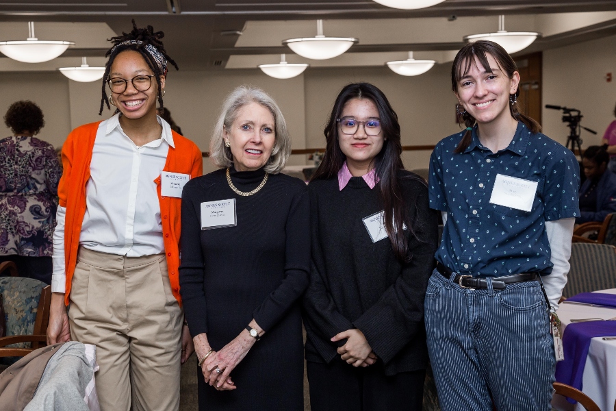 A group photo featuring four women. From left to right, they are standing and smiling, wearing name tags identifying them as participants in the Agnes Scott College event. The woman on the far left is wearing an orange cardigan and glasses, the next woman has short blonde hair and is dressed in black, the third woman is wearing glasses and a black outfit, and the woman on the far right has a blue polka dot shirt and light blue jeans.