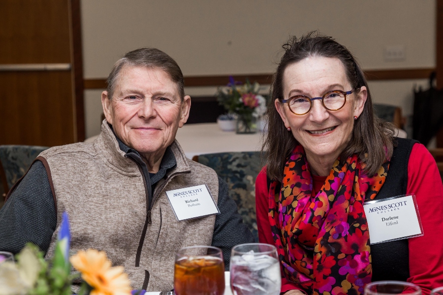 A close-up of two people sitting together at a dining table, both smiling. They are wearing name tags for Agnes Scott College. The man is wearing a beige zip-up vest over a black shirt, and the woman is wearing glasses, a colorful scarf, and a red top.