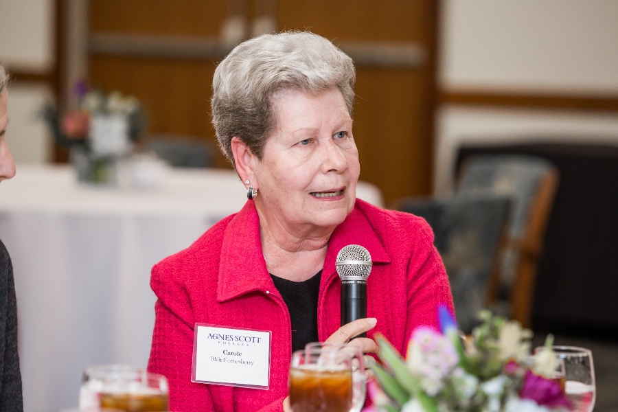 A woman holding a microphone while speaking at a dining table. She is wearing a bright pink jacket and a name tag for Agnes Scott College. She has short, gray hair and is seated in a room with other attendees in the background.