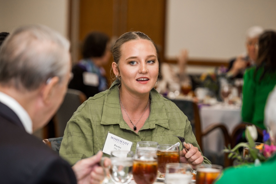 A young woman with blonde hair seated at a dining table, engaged in conversation. She is wearing a green button-up shirt and a name tag for Agnes Scott College. There are plates and glasses of iced tea on the table in front of her, and other attendees are visible in the background.